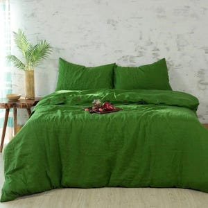 May include: A bed with green bedding, including a duvet cover and two pillowcases. A wooden side table holds a vase with green plants and a small bowl. A tray with fruit sits on the bed.