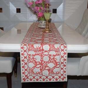 Può includere: Un runner da tavolo floreale rosso e bianco con un motivo a elefanti e fiori è esposto su un tavolo bianco. Il tavolo è circondato da sedie bianche. Un vaso con fiori rosa e un candelabro dorato sono sul tavolo.