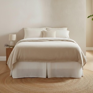 May include: A bed with a neutral color scheme, featuring a beige blanket and white bedding. The bed has a white bed skirt and is set on a round jute rug. A small lamp and side table are visible on the left.