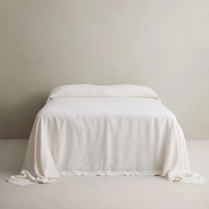 May include: A bed featuring a solid off-white bedspread that drapes over the sides and foot of the frame. The fabric has a soft, matte texture and covers the entire mattress surface, extending down to the floor with gentle folds at the corners.