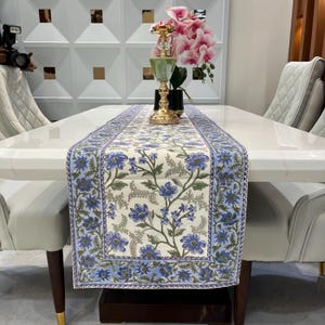 Può includere: Runner da tavolo floreale con sfondo crema e motivo floreale blu. Posizionato su un tavolo bianco, con un candelabro dorato e fiori rosa. Sedie bianche circondano il tavolo, creando un'atmosfera elegante.