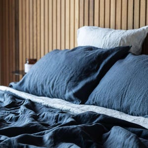 May include: A bed dressed with navy blue linen bedding, featuring two large pillows and a duvet cover. A light grey pillow is visible behind the navy pillows. The headboard is made of vertical wooden planks.