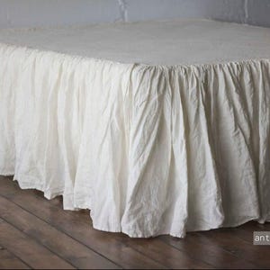 Pode incluir: Uma saia de cama branca antiga com um design franzido. O tecido é drapeado e texturizado, criando uma aparência macia e fluida. A saia de cama é mostrada em um piso de madeira.