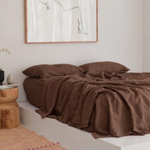 Peut inclure: Une parure de lit en lin marron foncé composée d'un drap plat et de deux taies d'oreiller assorties. Le tissu présente un tissage texturé visible et un aspect légèrement froissé. La literie est disposée sur un cadre de lit bas blanc.
