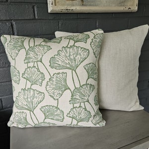 Op de afbeelding: Twee decoratieve kussens, een met een witte achtergrond en een groen ginkgo-bladpatroon, de andere effen lichtbeige.