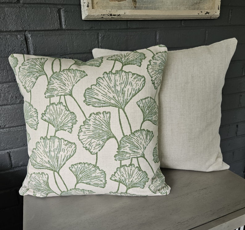 Op de afbeelding: Twee decoratieve kussens, een met een witte achtergrond en een groen ginkgo-bladpatroon, de andere effen lichtbeige.