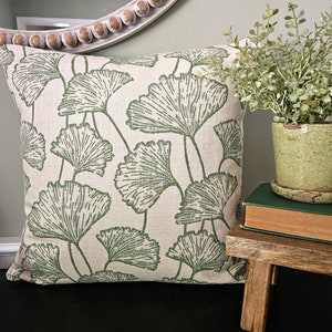 Op de afbeelding: Een cr&egrave;mekleurig kussen met een groen ginkgo-bladpatroon. Het kussen ligt op een zwart oppervlak naast een kleine houten kruk met een boek en een potplant.