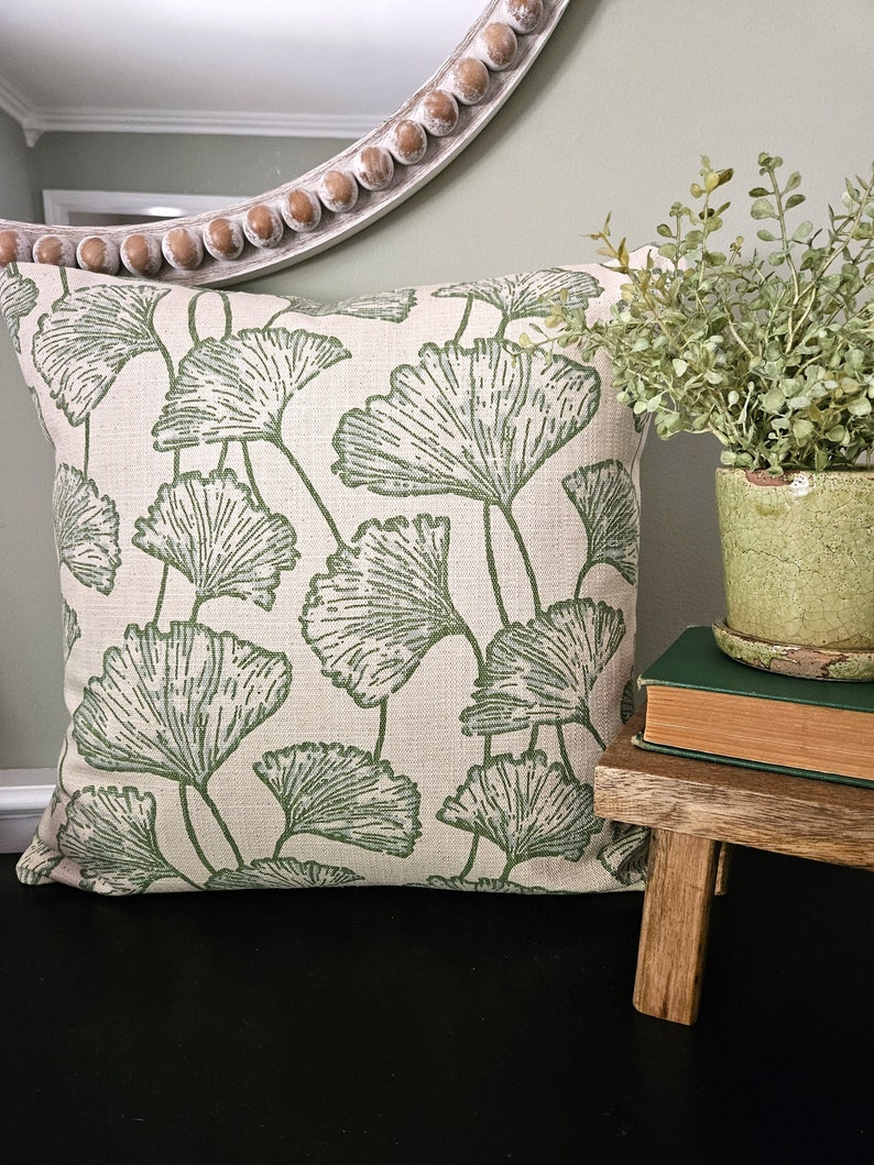 Op de afbeelding: Een cr&egrave;mekleurig kussen met een groen ginkgo-bladpatroon. Het kussen ligt op een zwart oppervlak naast een kleine houten kruk met een boek en een potplant.