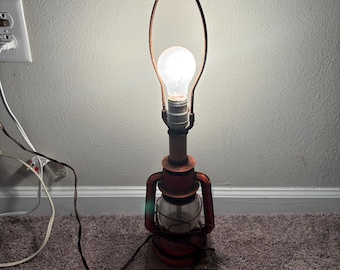 Può includere: Una lampada in stile antico con una base a lanterna in metallo rosso e un globo in vetro trasparente. La lampada ha una lampadina accesa e un telaio color rame. La lampada è collegata.