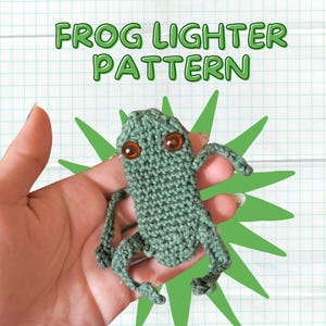 May include: A crocheted frog lighter pattern is displayed. The frog is green with orange eyes and is held in a hand. The text "FROG LIGHTER PATTERN" is in green letters above the frog, with a green starburst design behind it.