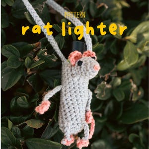 Może przedstawiać: Szydełkowy pokrowiec na zapalniczkę w kształcie szczura, w kolorze szarym z różowymi akcentami. Szczur ma czarny cylinder i wisi na szarym sznurku. Tekst "rat lighter" jest w kolorze żółtym, z napisem "pattern" nad nim.