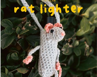 Puede incluir: Funda de encendedor de ratón de ganchillo en gris con detalles rosas. El ratón tiene un sombrero de copa negro y cuelga de un cordón gris. El texto "rat lighter" está en amarillo, con la palabra "pattern" encima.