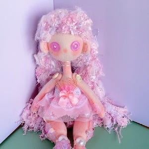 Può includere: Bambola di stoffa fatta a mano con capelli rosa e un vestito rosa. La bambola ha grandi occhi rotondi ed è adornata con paillettes, perle e abbellimenti floreali. Le gambe della bambola sono decorate con fiori e paillettes. La bambola è ambientata su uno sfondo verde e viola.
