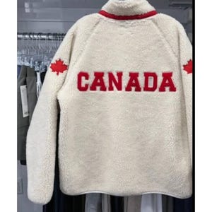 May include: Cream-colored fleece jacket with the word "CANADA" in red block letters across the back. Features a red maple leaf on each shoulder and a red trim around the collar.