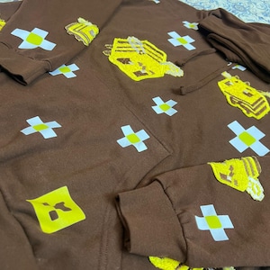 K&ouml;nnte beinhalten: Brauner Kapuzenpullover mit gestickten gelben und wei&szlig;en Designs. Der Hoodie hat ein sich wiederholendes Muster aus wei&szlig;en Kreuzen mit gelben Zentren und mehreren gr&ouml;&szlig;eren gelb gestickten Formen. Die &Auml;rmel und die Kapuze sind sichtbar.