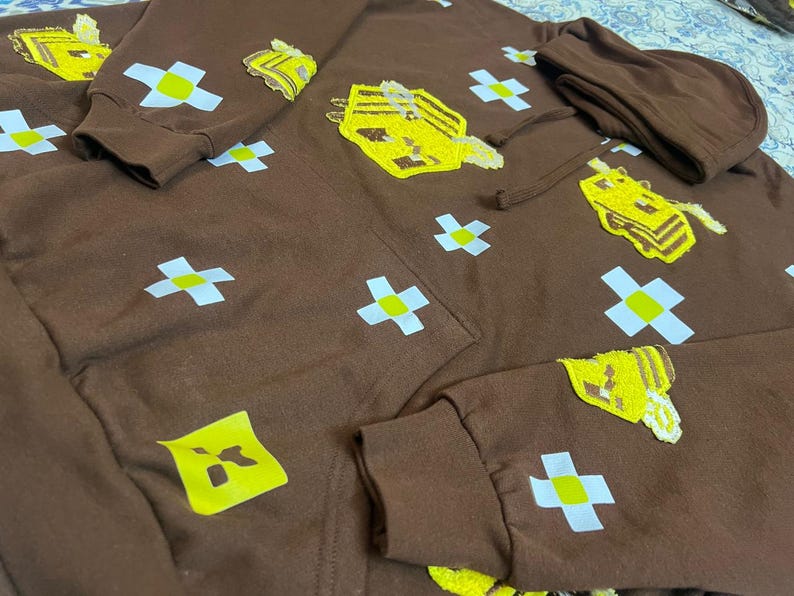 K&ouml;nnte beinhalten: Brauner Kapuzenpullover mit gestickten gelben und wei&szlig;en Designs. Der Hoodie hat ein sich wiederholendes Muster aus wei&szlig;en Kreuzen mit gelben Zentren und mehreren gr&ouml;&szlig;eren gelb gestickten Formen. Die &Auml;rmel und die Kapuze sind sichtbar.