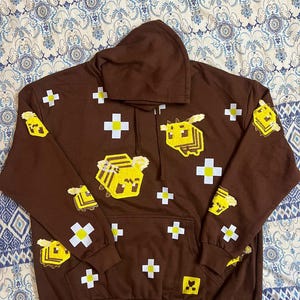 K&ouml;nnte beinhalten: Ein brauner Kapuzenpullover mit einer Vordertasche und einer Kapuze mit Kordelzug. Der Pullover zeigt pixelige gelbe Bienen und wei&szlig;e Pluszeichen. Die Bienen befinden sich auf der Vorderseite, den &Auml;rmeln und der Kapuze.