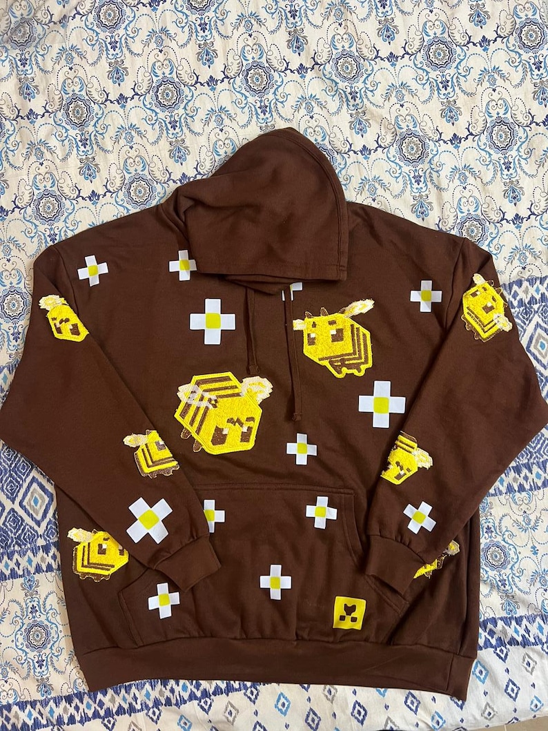 K&ouml;nnte beinhalten: Ein brauner Kapuzenpullover mit einer Vordertasche und einer Kapuze mit Kordelzug. Der Pullover zeigt pixelige gelbe Bienen und wei&szlig;e Pluszeichen. Die Bienen befinden sich auf der Vorderseite, den &Auml;rmeln und der Kapuze.