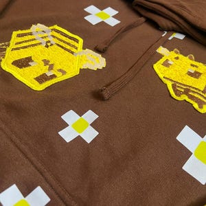 K&ouml;nnte beinhalten: Brauner Hoodie mit gestickten gelben und braunen Bienen- und Mausmotiven. Der Hoodie hat auch wei&szlig;e und gelbe kreuzf&ouml;rmige Akzente. Der Kordelzug und die Kapuze sind im Bild zu sehen.