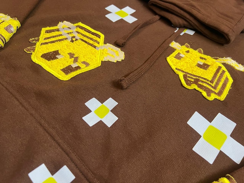 K&ouml;nnte beinhalten: Brauner Hoodie mit gestickten gelben und braunen Bienen- und Mausmotiven. Der Hoodie hat auch wei&szlig;e und gelbe kreuzf&ouml;rmige Akzente. Der Kordelzug und die Kapuze sind im Bild zu sehen.