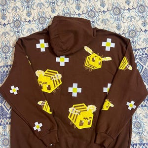 K&ouml;nnte beinhalten: Ein brauner Kapuzenpullover mit pixeligen gelben Bienen und wei&szlig;en Kreuzen. Das Design wiederholt sich auf der Vorderseite und den &Auml;rmeln. Der Hoodie hat einen Kordelzug und eine K&auml;ngurutasche.