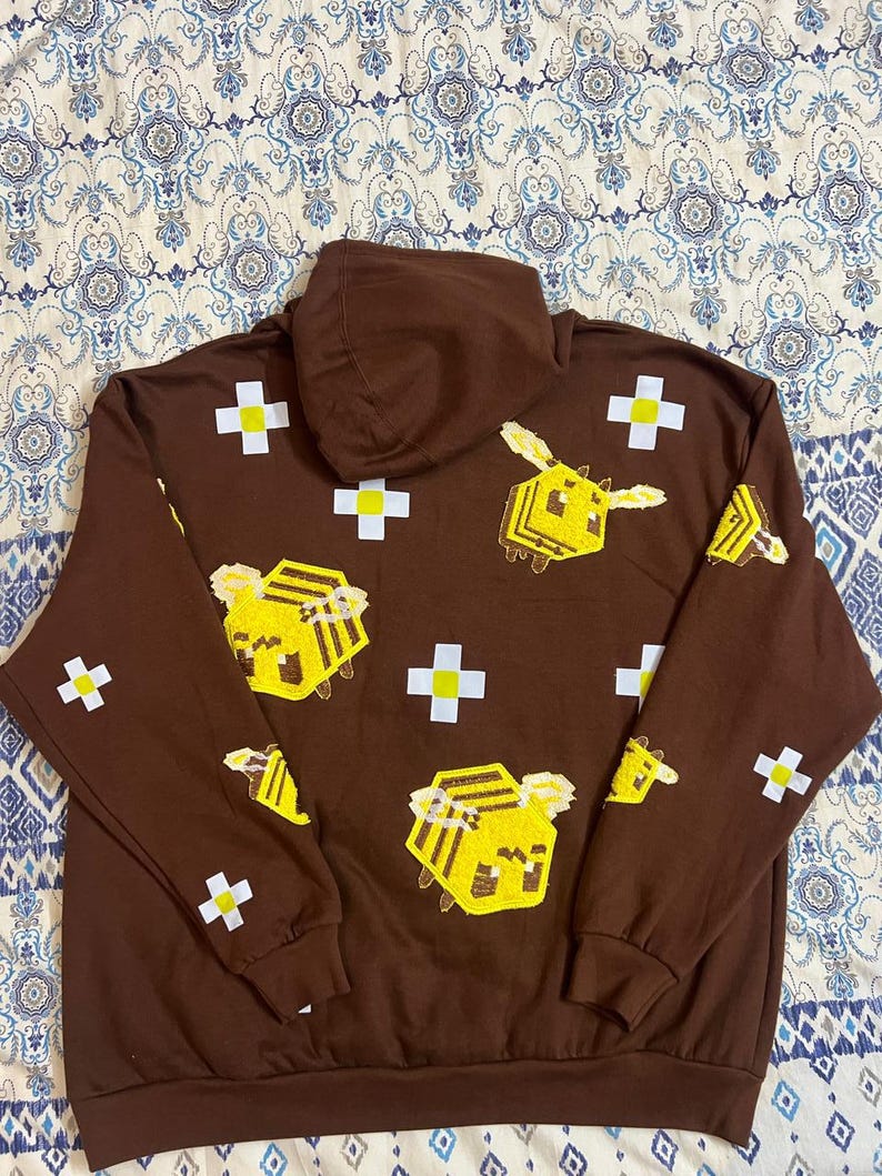 K&ouml;nnte beinhalten: Ein brauner Kapuzenpullover mit pixeligen gelben Bienen und wei&szlig;en Kreuzen. Das Design wiederholt sich auf der Vorderseite und den &Auml;rmeln. Der Hoodie hat einen Kordelzug und eine K&auml;ngurutasche.
