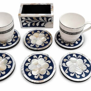 May include: A set featuring two white ceramic mugs with dark patterned bands, a rectangular holder, and five circular coasters, all decorated with dark blue surfaces and white floral mother-of-pearl inlay, accompanied by five small polished stones.