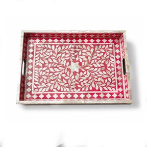 Peut inclure: Un plateau de service rectangulaire avec un fond rose vif, orné d'un motif complexe de feuilles blanches et d'une étoile centrale. La bordure présente un design en losanges répétés et les bords sont finis avec une incrustation claire.