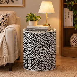 May include: A cylindrical side table featuring a black and white leaf pattern. The table is topped with two stacked books titled The Kinfolk Home and At Home, a small potted green plant, and a brass-toned lamp with a pleated white fabric shade.