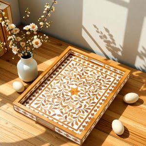 May include: A rectangular wooden serving tray featuring a white inlay with a brown leaf and vine pattern. The tray has integrated handles on the short sides and a border of white floral motifs. It is positioned near a white vase with white flowers.