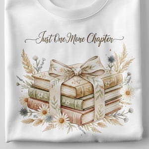 May include: White t-shirt featuring a watercolor design of a stack of books tied with a bow, surrounded by flowers and foliage. The text "Just One More Chapter" is written above the books in a script font. A pen and notebook are on the left.