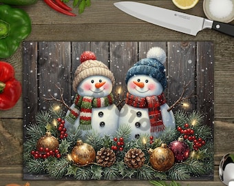 Könnte beinhalten: Rechteckiges Schneidebrett mit zwei Schneemännern, die Hüte und Schals tragen, umgeben von Weihnachtsschmuck. Die Schneemänner haben Karottennasen und Kohleaugen. Der Hintergrund ist ein Holzzaun.
