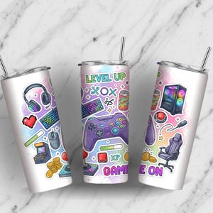 May include: Three white stainless steel tumblers with gaming-themed stickers. Each tumbler features a silver straw and colorful graphics including a controller, keyboard, headphones, and the text "LEVEL UP" and "GAME ON".