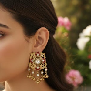May include: Gold-coloured, elaborate earrings with a square top, red and white details. The earrings have a central clear stone and dangling teardrop charms. The design is intricate and detailed, with a vintage aesthetic.
