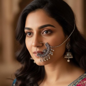 May include: A silver nose ring and matching earrings. The nose ring has a crescent design with dangling beads. The earrings are bell-shaped with a pearl at the bottom. The jewelry is worn with a teal and pink embroidered top.