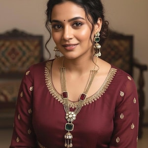 May include: A woman wearing a maroon dress with gold embroidery and a multi-layered gold necklace with red and green gemstone accents. Matching earrings complete the look. The dress has long sleeves and a detailed neckline.