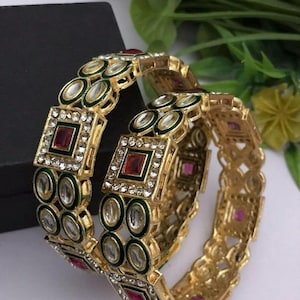 May include: Gold-toned bangles with intricate detailing. The bangles feature square and oval-shaped accents with red and clear gemstones, and green enamel. The design is ornate and eye-catching, suitable for special occasions.