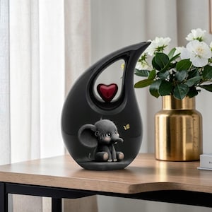 Puede incluir: Una urna negra en forma de lágrima con un acento rojo en forma de corazón. La urna presenta una ilustración de un elefante gris sentado con orejas grandes y una pequeña mariposa. A la derecha hay un jarrón dorado con flores blancas.
