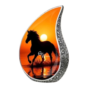 Op de afbeelding: Een druppelvormige hanger met een zonsondergangscène met een zwart paardensilhouet dat door water rent. De achtergrond is een levendige oranje en gele gradiënt. De hanger heeft een zilverkleurige, sierlijke rand met de tekst "Crafted Whispers".
