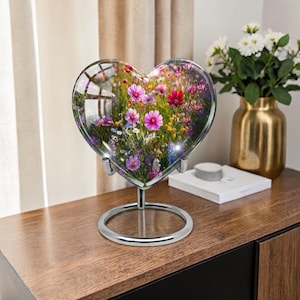 May include: A heart-shaped glass sculpture filled with colourful flowers, mounted on a silver stand. The flowers include pink, purple, and red blooms. The sculpture sits on a wooden surface, with a gold vase and white flowers in the background.