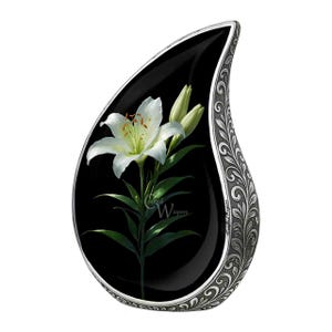 May include: A teardrop-shaped memorial urn with a black background featuring a white lily flower and green leaves. The urn has a silver-colored, ornate, floral design around the edges. The text "crafted whispers" is visible.