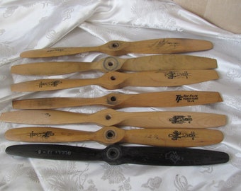 May include: Eight wooden airplane propellers with black lettering and a single black propeller. The propellers are arranged in a row on a white fabric background. The propellers have various markings, including "Top Flight Power Plant" and "9-E7 17708".