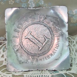 May include: A silver metal ashtray with a raised design of a lion's head and the words "Lions International" around a circle with a large "L" in the center.