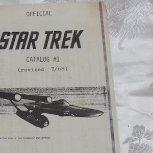 May include: A black and white image of the Starship Enterprise from the original Star Trek series. The image is from a catalog for Star Trek merchandise. The text reads "OFFICIAL STAR TREK CATALOG #1 (revised 7/68) FROM THE LOG OF THE STARSHIP ENTERPRISE YOUR PASSPORT TO A GALAXY OF AUTHENTIC STAR TREK COLLECTOR'S ITEMS!"