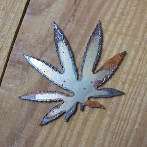 May include: A metal cannabis leaf with a rustic, aged finish. The leaf is a light silver colour with brown rust spots.