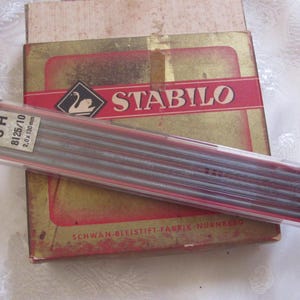 May include: A package of Stabilo 3H pencil leads, 2.0 x 130 mm, rests on a vintage box with the Stabilo logo. The box has a red banner with the brand name and a swan emblem. The packaging is aged with a gold and red color scheme.
