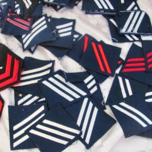 May include: A selection of vintage navy blue and black military patches, featuring white and red stripes and insignia. The patches are displayed on a white fabric, showcasing various rank and service emblems.