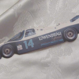 May include: A vintage race car toy in blue and white, featuring the number 14 and the "Löwenbräu Special" logo. The car has Goodyear tires and a Porsche logo, suggesting a collectible item.