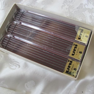 May include: A box of Uni brand B leads for mechanical pencils. The box contains three clear plastic tubes filled with dark red leads. Each tube is labeled with the Uni logo and "6 Leads B". The box is beige.