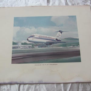 May include: A vintage print of a white Douglas DC-9 jet transport airplane with red and blue accents. The plane is on a runway with a blue sky and white clouds in the background. The text "DOUGLAS DC-9 JET TRANSPORT" is printed below the plane.